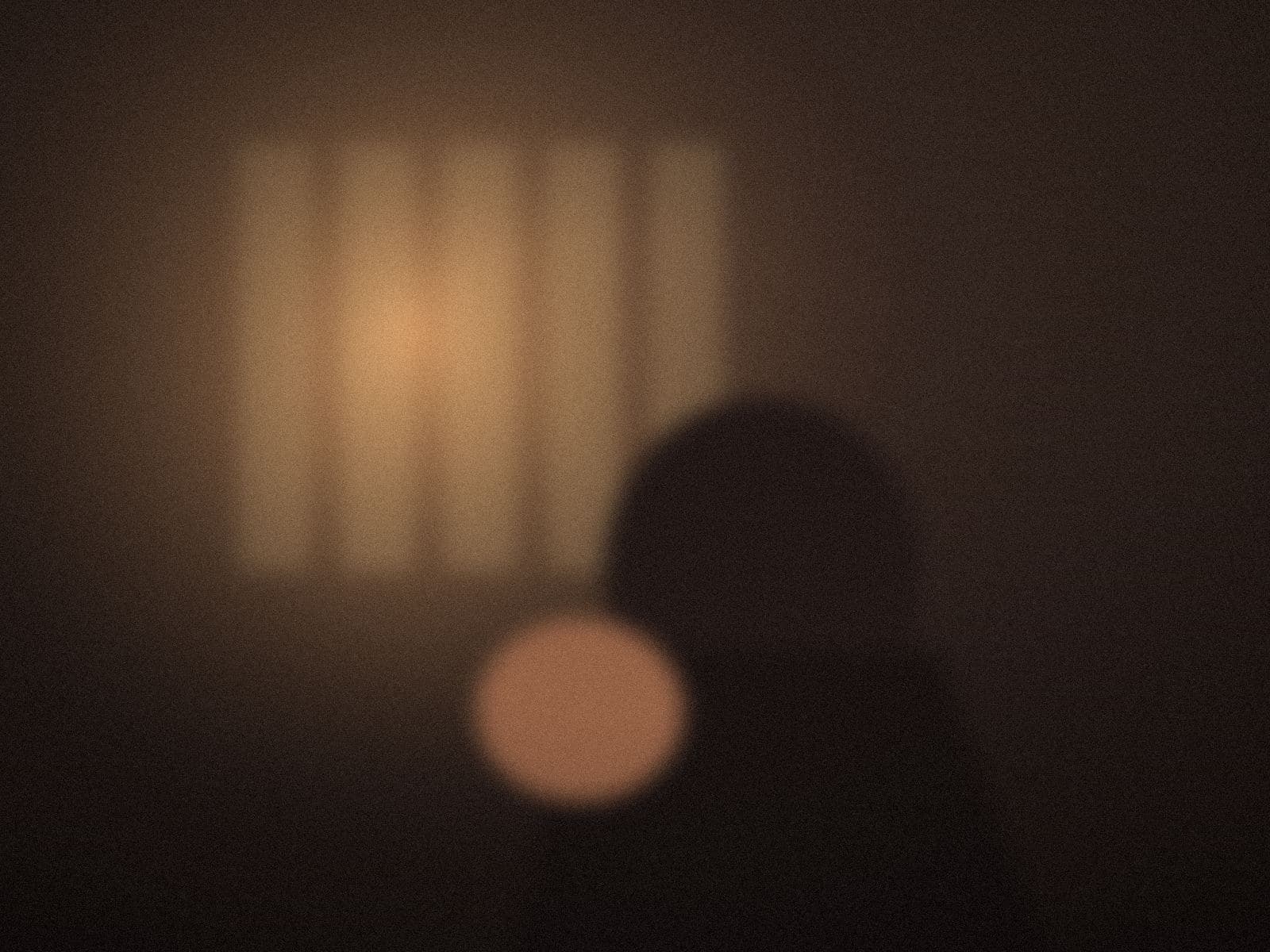 A figure in a dim room beside a window, striped light on the wall